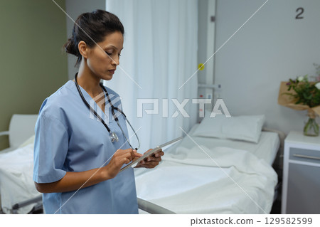 Mature nurse wearing blue scrubs and stethoscope standing beside bed in patient room holding tablet 129582599