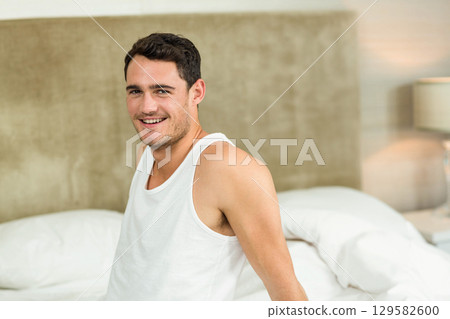 Male sitting on bed smiling under lamp with white sheets and beige headboard leaning back Male sitting on bed smiling under lamp with white sheets and beige headboard leaning back 129582600