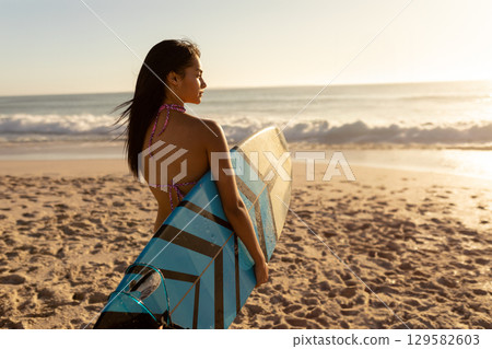Asian woman in her twenties wearing bikini holding blue-and-black striped surfboard at water edge 129582603