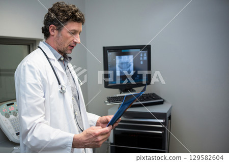 Middle-aged doctor holding blue x-ray folder with stethoscope and examining monitor in clinic 129582604