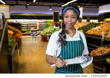 African American woman wearing striped apron checking produce with clipboard in supermarket aisle African American woman wearing striped apron checking produce with clipboard in supermarket aisle 129582605