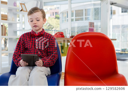 Preschool boy sitting on blue chair holding tablet in lounge beside rotary telephone, copy space 129582606