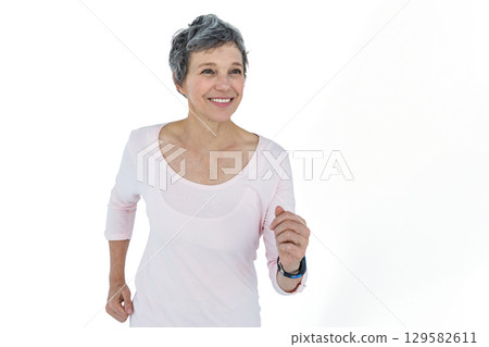 Senior female running in place on lit white backdrop wearing pink workout top, fitness tracker 129582611