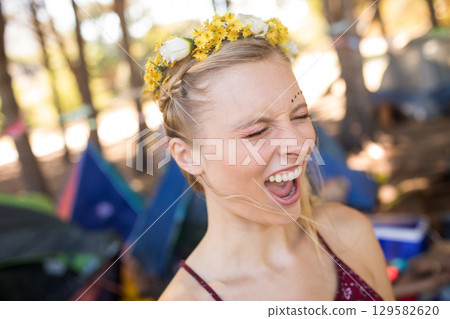 Woman standing among tents at campsite wearing burgundy spaghetti strap top, flower crown and gems 129582620