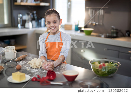 Female child kneading dough in orange apron on kitchen counter with flour jar and rolling pin 129582622