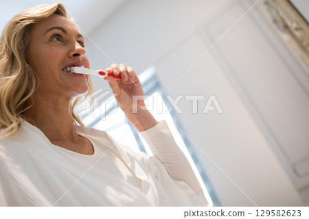 Mid adult woman brushing teeth with red white toothbrush in bathroom wearing bathrobe 129582623