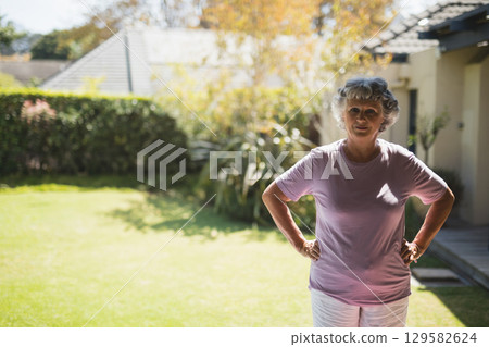 Senior woman standing with hands on hips in suburban backyard by hedge and deck, copy space 129582624