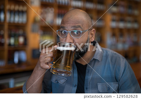 Indian man wearing eyeglasses lifting clear glass mug with beer foam while sitting at pub counter 129582628