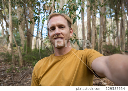 Middle-aged man holding smartphone and taking selfie in eucalyptus forest wearing mustard T-shirt 129582634