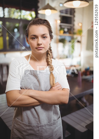 Woman standing with folded arms in cafe wearing grey apron near wooden tables and pendant lights Woman standing with folded arms in cafe wearing grey apron near wooden tables and pendant lights 129582639