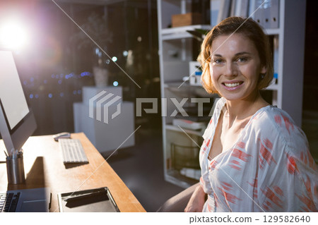 Woman smiling, working on drawing tablet with stylus on desk under lamp in office at night 129582640