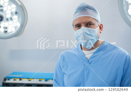 Senior male surgeon standing in scrubs in operating room under surgical lights by instrument table Senior male surgeon standing in scrubs in operating room under surgical lights by instrument table 129582649