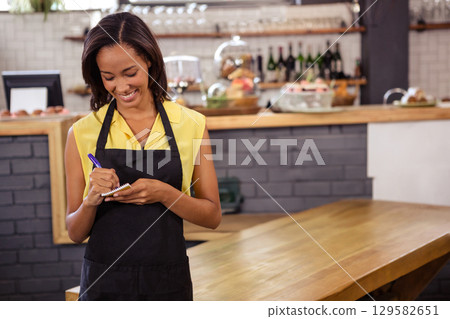 African American woman in apron standing behind cafe counter writing on notepad beside pastries 129582651