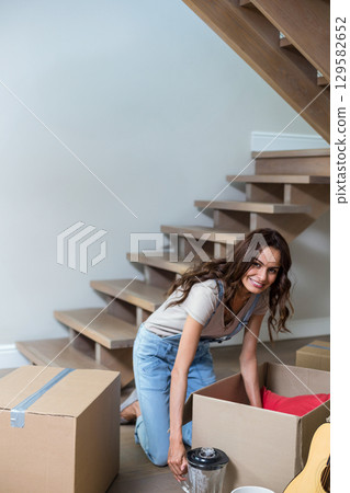 Mid adult woman kneeling on floor unpacking arranging boxes blender pitcher and guitar, copy space Mid adult woman kneeling on floor unpacking arranging boxes blender pitcher and guitar, copy space 129582652