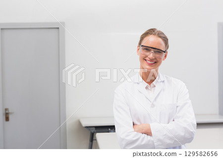 Female scientist smiling while standing in clean lab, wearing lab coat and safety goggles 129582656