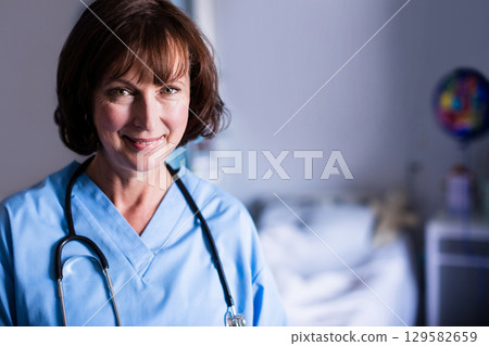 Smiling female nurse standing in patient room in blue scrubs with stethoscope, copy space 129582659
