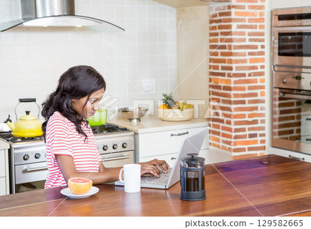 African American woman wearing red striped top working on laptop at kitchen island with coffee mug 129582665