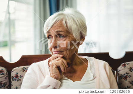 Senior woman sitting pensively on floral couch with cushions in living room with sheer curtains 129582666