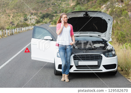 Woman leaning against sedan hood open on mountain road placing red triangle calling on smartphone 129582670
