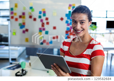 Mid-20s woman smiling and holding tablet in office by whiteboard with sticky notes, copy space Mid-20s woman smiling and holding tablet in office by whiteboard with sticky notes, copy space 129582684