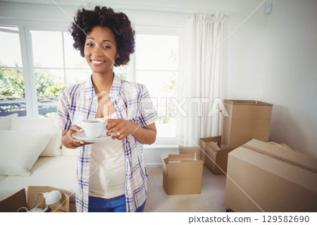 African American woman standing amid moving boxes near sofa in living room holding cup and saucer 129582690