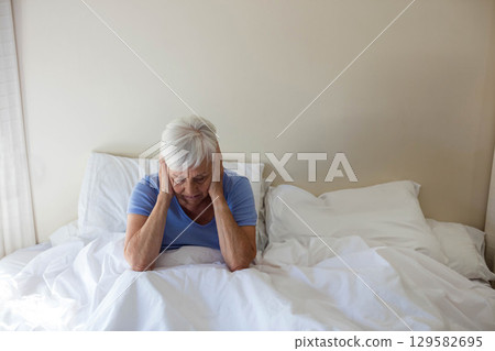 Senior woman sitting on bed wearing light blue shirt holding hands to temples on white comforter Senior woman sitting on bed wearing light blue shirt holding hands to temples on white comforter 129582695