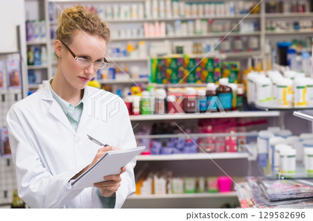 Female pharmacist writing on notepad with pen at pharmacy counter wearing lab coat, copy space 129582696