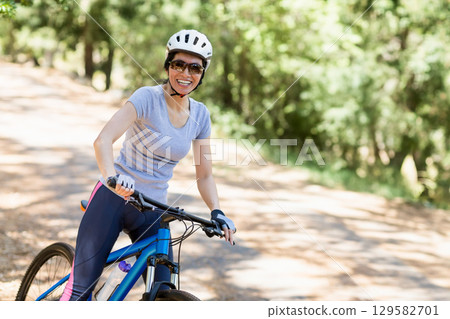Female cyclist riding blue bicycle along sunlit dirt trail in woods wearing helmet and sunglasses Female cyclist riding blue bicycle along sunlit dirt trail in woods wearing helmet and sunglasses 129582701