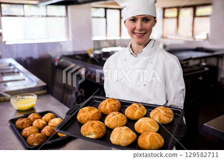 Female chef in chef coat and hat, holding baking tray with golden rolls in restaurant kitchen 129582711