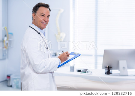 Smiling male doctor wearing stethoscope and holding blue clipboard in clinic exam room, copy space 129582716