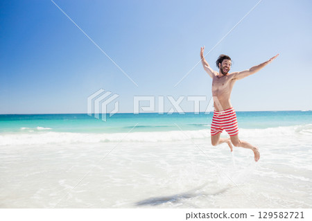 Male jumping in shallow shoreline water wearing red and white striped swim trunks, copy space 129582721
