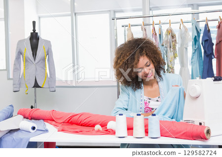 Woman with curly hair sewing red fabric on sewing machine at fashion studio with measuring tape Woman with curly hair sewing red fabric on sewing machine at fashion studio with measuring tape 129582724