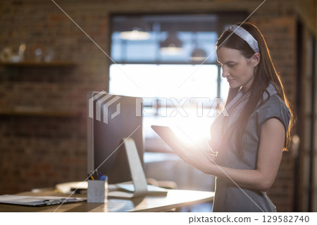 Female professional in dress standing at desk in office reviewing tablet and monitor, copy space 129582740