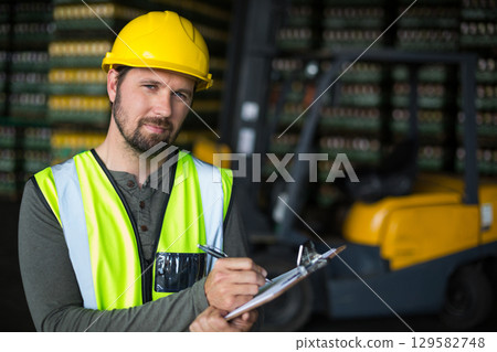 Middle-aged man inspecting warehouse inventory with clipboard wearing hard hat and vest, copy space Middle-aged man inspecting warehouse inventory with clipboard wearing hard hat and vest, copy space 129582748