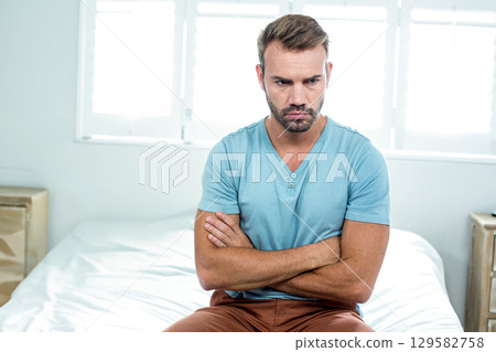 Mid-adult man sitting on bed edge in bright bedroom crossing arms near nightstand, box and blinds Mid-adult man sitting on bed edge in bright bedroom crossing arms near nightstand, box and blinds 129582758