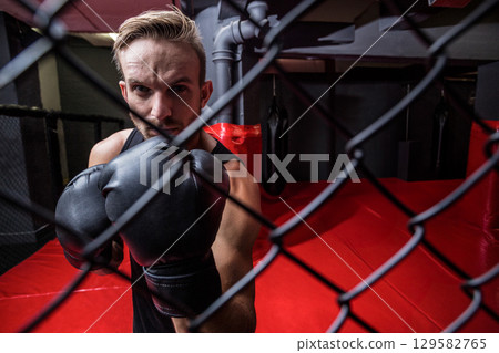 Male athlete holding boxing gloves up inside chain-link fight cage at gym with red floor 129582765