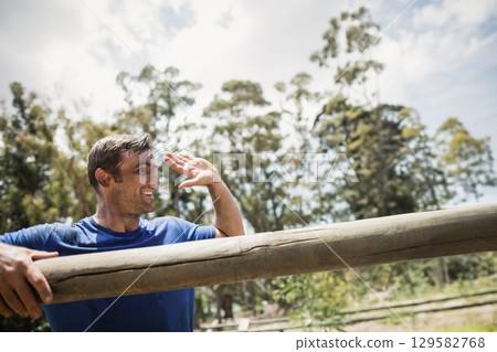 Middle-aged man leaning on wooden beam at obstacle course, shielding eyes in blue shirt, copy space 129582768