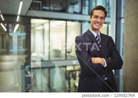 Man in suit leaning arms crossed against column at office atrium with glass railing, copy space 129582769