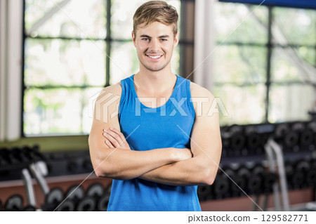 Man standing with arms crossed in gym area wearing blue sleeveless top with dumbbells and windows Man standing with arms crossed in gym area wearing blue sleeveless top with dumbbells and windows 129582771