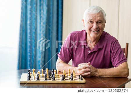Senior man sitting at wooden table at home studying chessboard setup near blue patterned curtains 129582774