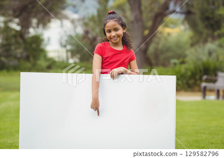 School-age girl wearing red t-shirt and jeans standing on park lawn pointing at blank white sign School-age girl wearing red t-shirt and jeans standing on park lawn pointing at blank white sign 129582796