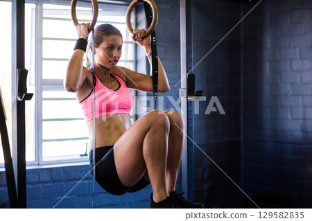 Fit woman wearing pink sports bra, black shorts holding wooden rings and lifting knees at gym 129582835
