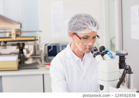 Woman scientist in lab coat examining sample under microscope at lab bench with computer monitor 129582836