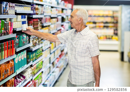 Senior male shopper grabbing carton from supermarket shelving near fridge display, copy space 129582856
