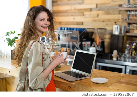 Woman standing at coffee shop bar holding white cup with silver laptop, copy space Woman standing at coffee shop bar holding white cup with silver laptop, copy space 129582867