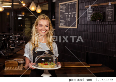 Female barista wearing apron standing behind espresso machine at cafe holding tart under glass dome Female barista wearing apron standing behind espresso machine at cafe holding tart under glass dome 129582876