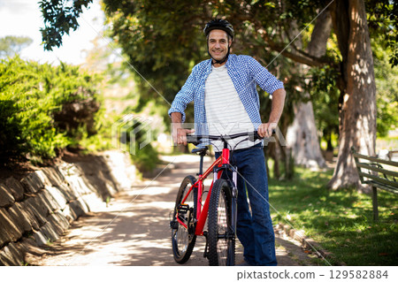 Man wearing helmet pushing red bicycle along sunlit park path with stone wall and park bench 129582884
