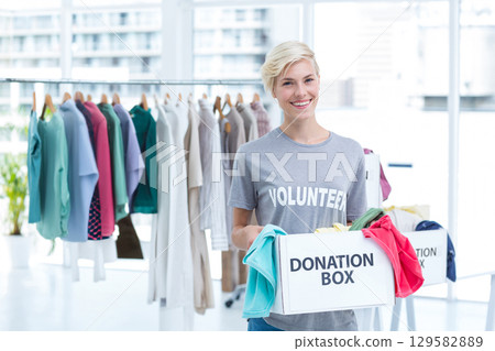 Smiling female volunteer holding donation box and standing by clothing rack in donation center 129582889