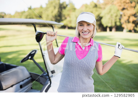 Senior woman holding driver standing on fairway by golf cart wearing golf attire, visor and glove 129582905