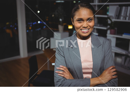 Businesswoman standing in modern office at night with desk, shelves with plant and city skyline 129582906